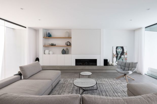 Bright, spacious contemporary living room with a neutral grey and white palette. A large sectional sofa surrounds layered round coffee tables and a textured area rug, facing a built-in media wall with open display shelving, white cabinetry, a linear fireplace, and vertical fluted wall panelling. Full-height glazing with sheer curtains and a sculptural lounge chair complete the refined minimalist setting.