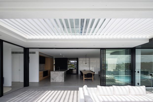 A bright, contemporary open-plan interior with a kitchen, dining area and living space opening onto a terrace or pool edge. Full-height black-framed glazing and sliding doors create a strong indoor-outdoor connection, while a white slatted ceiling and matching patterned shadows add a distinctive architectural feature. The kitchen includes a dark island with a pale stone benchtop, and a white sofa occupies the foreground.