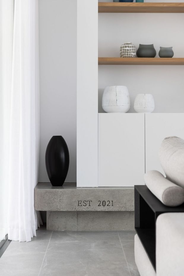 A close-up view of a refined contemporary living room featuring a pale grey tiled floor, white sheer curtains, a built-in white storage unit with open timber shelves, and minimalist ceramic vessels. A black sculptural vase sits on a substantial concrete console marked “EST 2021,” while the edge of a cream upholstered sofa and black frame is visible in the foreground. The palette is restrained in white, grey, black, and natural timber tones.