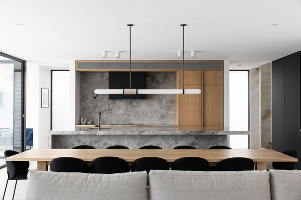 Contemporary open-plan kitchen and dining area viewed from behind a large light-upholstered sofa. The kitchen features a long stone island with integrated seating, a textured grey stone backsplash, warm timber cabinetry, dark built-in cabinetry, and slim linear pendant lights. A large timber dining table with black chairs occupies the foreground, with bright white walls and expansive glazing contributing to an airy, refined atmosphere.