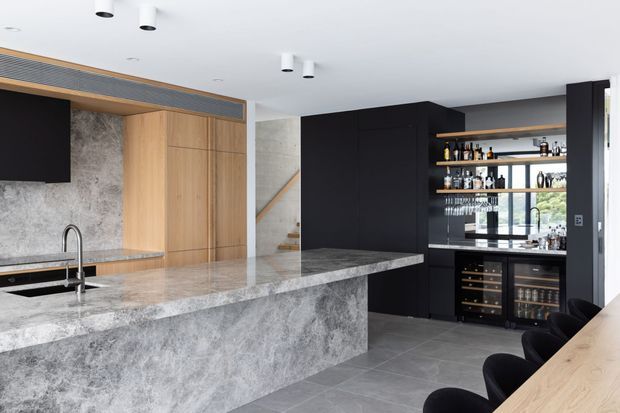 Contemporary open-plan kitchen and home bar featuring a large grey stone island with integrated sink, dark cabinetry, timber veneer storage, open bottle shelving, and built-in wine and beverage fridges. Grey tiled flooring, white ceiling, and a partially visible stairway complete the refined, monochromatic interior.