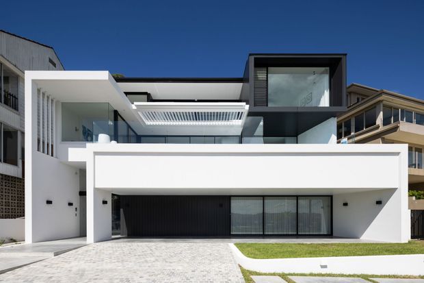 Street-facing view of a contemporary multi-storey residence with a crisp white rendered facade, strong horizontal and cubic forms, extensive glazing, a dark recessed garage, rooftop terrace elements, and a paved driveway with a narrow lawn strip. Nearby houses are visible at the edges.
