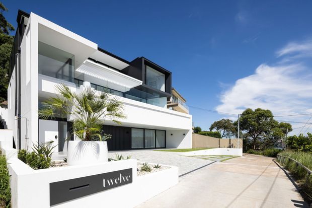 Street-facing view of a contemporary multi-level residence with a crisp white rendered facade, strong geometric forms, dark framed glazing, recessed balconies, and a flat roofline. The property includes landscaped raised planters, palm planting, a broad concrete driveway, and a partially visible rooftop or upper-level terrace.