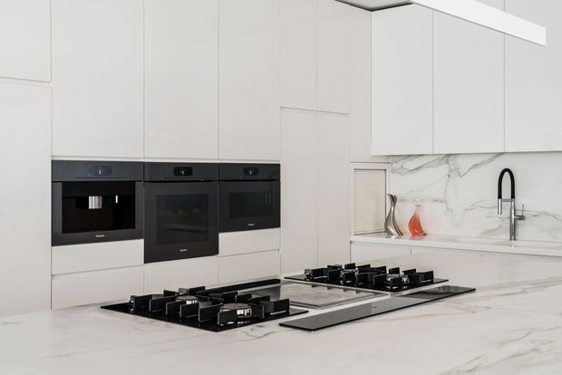 A bright, refined contemporary kitchen with extensive white full-height cabinetry, integrated black appliances, a large white marble-look island, and two black gas cooktops. The rear wall features a matching marble-look splashback and a black gooseneck tap at the sink. Minimal decorative styling creates a clean, high-end appearance.