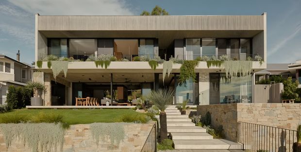 A contemporary multi-level residential house viewed from the front, featuring a flat-roofed rectangular form, extensive dark-framed glazing, pale textured exterior walls, deep covered outdoor living areas, and planted balconies with cascading greenery. Broad steps lead through a landscaped garden toward the entrance, with pale stone retaining walls and metal handrails. Neighbouring houses are visible on both sides.