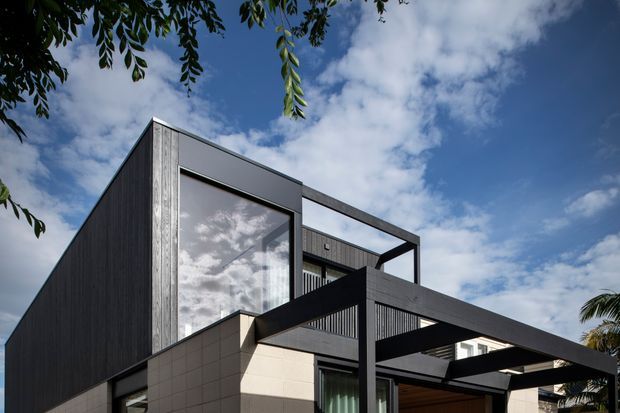 Exterior architectural photograph of a contemporary multi-level house with sharply geometric massing, dark vertical cladding, pale tiled or panelled lower walls, extensive glazing, and a projecting black-framed pergola or carport structure. The image is viewed from below against a partly cloudy blue sky, with some tree foliage framing the composition.