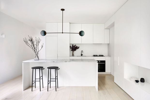 A bright, minimalist white kitchen with a large marble-look island, two black bar stools, white flat-panel cabinetry, integrated appliances, a compact sink, and pale timber flooring. A sculptural black pendant light and sparse branch arrangement provide restrained accents. The room appears compact to medium-sized with a clean, calm contemporary feel.