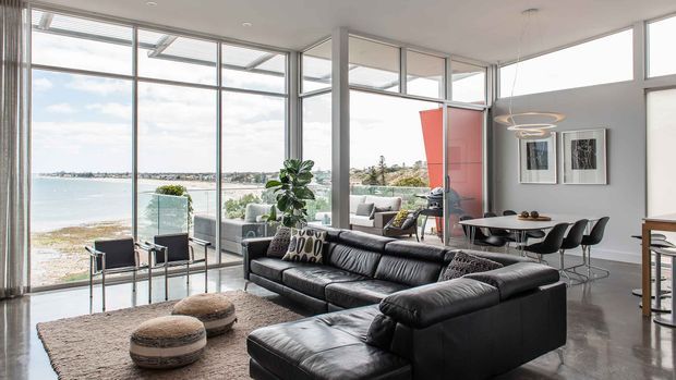 Bright, open-plan contemporary living area with a large black sectional sofa, textured neutral rug and low poufs, opening directly to a dining zone. Floor-to-ceiling glazing provides expansive waterfront views, while a sculptural ring pendant, framed wall art, indoor planting and a red vertical feature panel add visual interest.
