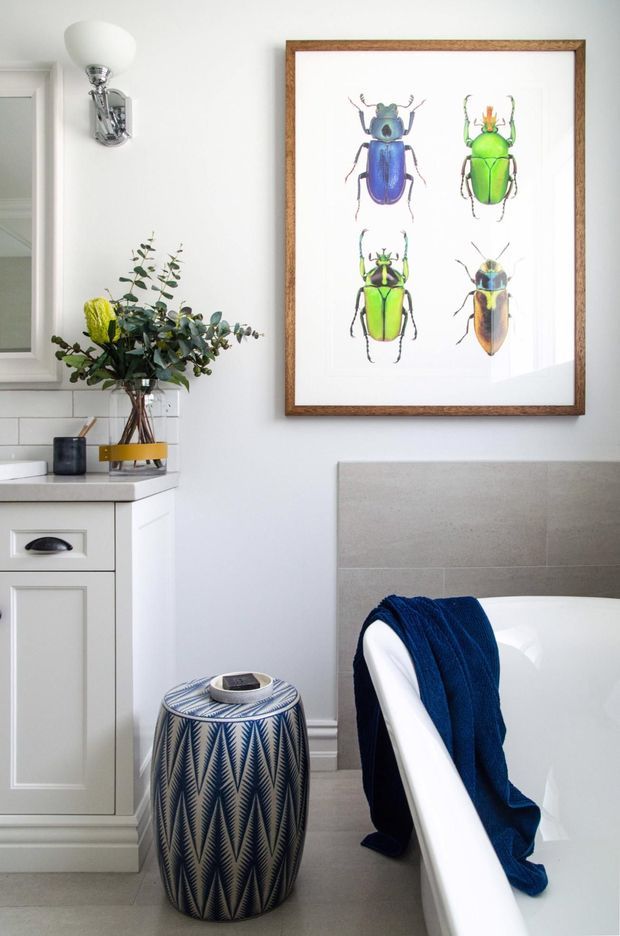 A bright, refined bathroom featuring a freestanding white bathtub, pale grey tiled wall, white vanity cabinetry with a light benchtop, chrome wall sconce, and a framed botanical beetle print. A patterned blue-and-white stool and dark blue towel add colour and texture.