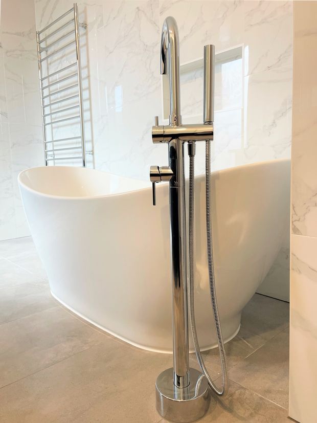 Bright contemporary bathroom featuring a freestanding white bathtub, polished chrome floor-mounted bath and shower fixture, chrome heated towel rail, large-format pale marble-look wall tiles, and light grey floor tiles. Natural light enters through a window behind the tub.