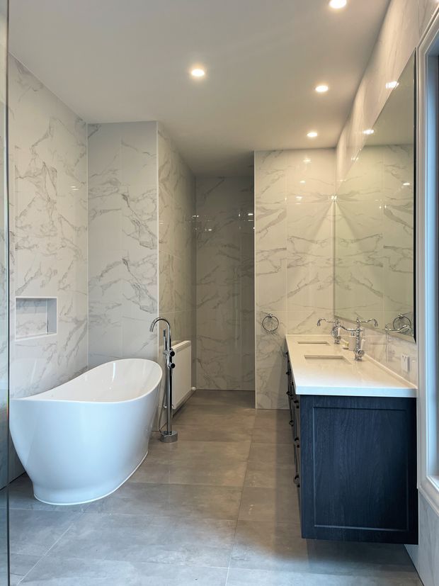 A bright, contemporary bathroom featuring a freestanding white bathtub, a double vanity with dark blue cabinetry and white countertop, large-format white marble-look wall tiles, grey floor tiles, recessed ceiling lights, and a walk-in shower at the rear.