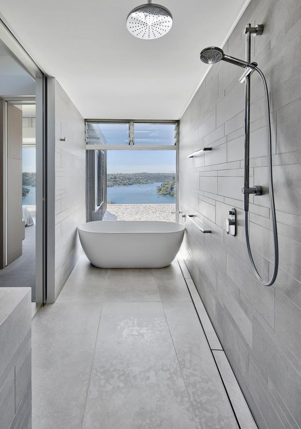 A narrow, contemporary bathroom with a freestanding white bathtub positioned beside a large waterside window. The room features pale grey tiled walls and floor, a linear floor drain, chrome wall-mounted shower hardware with a rain shower, recessed-style wall lighting, and a frameless glass opening toward an adjoining bedroom. The view beyond the window shows a bay and hillside.