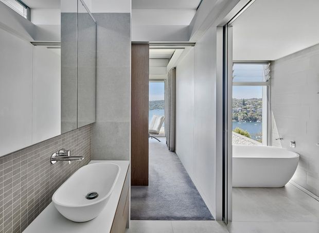 A bright, contemporary bathroom with a freestanding white bathtub in the foreground, wall-mounted chrome tapware, grey small-format tiled splashback, and a pale stone or tiled vanity surface. A second bathroom with another freestanding tub is visible through the open doorway on the right, while a carpeted corridor leads toward a sunlit sitting area and water view. The space has clean white walls, concealed or minimal detailing, large glazing, and a calm, refined finish.