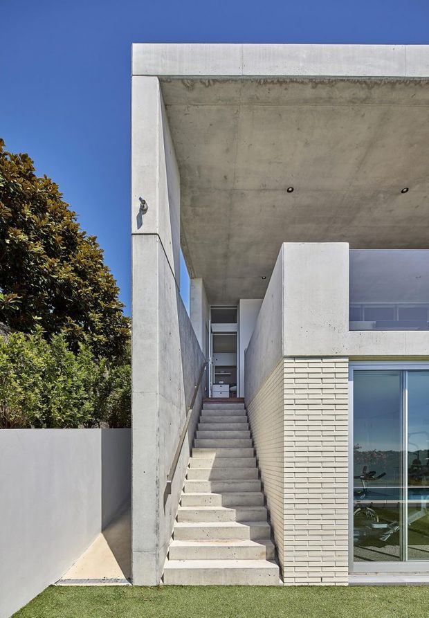 Exterior architectural photograph of a contemporary multi-level residence with a strong exposed-concrete frame and deep cantilevered roof or balcony slab. A straight concrete stairway rises between a tall concrete wall and a pale brick-clad volume toward an upper entrance. Large glazed doors or windows are visible at ground level, with a manicured lawn and dense tree at the edge of the property.
