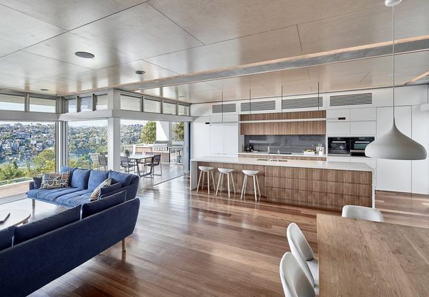 A bright, open-plan contemporary kitchen, dining, and living area with expansive corner glazing overlooking a leafy urban landscape. The kitchen features white full-height joinery, a timber island with integrated seating, stainless-steel appliances, and a timber-lined open shelf. Rich timber flooring continues through the space, with a navy sectional sofa, timber dining table, white dining chairs, and a large sculptural pendant light. The ceiling includes recessed services and linear ventilation details.