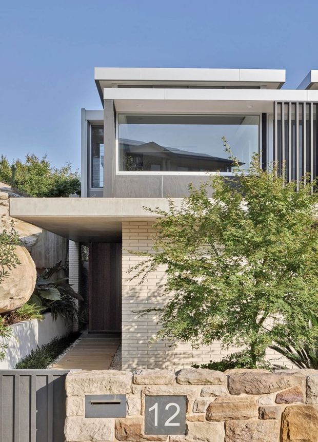 Contemporary residential facade viewed from the street, featuring layered flat roof planes, a projecting concrete canopy, large horizontal glazing, vertical metal screening, pale brickwork, and a rough natural-stone boundary wall. Dense planting partially obscures the lower facade and entry.