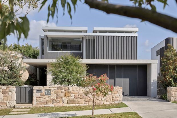 Contemporary multi-storey residence viewed from the street, with a flat roof, projecting concrete slab, extensive glazing, and a prominent upper-storey vertical louvred screen. The facade combines smooth light-grey concrete, dark garage doors and frames, and warm stacked stone retaining walls. A paved driveway, front path, low landscaping, and neighbouring modern homes are visible.