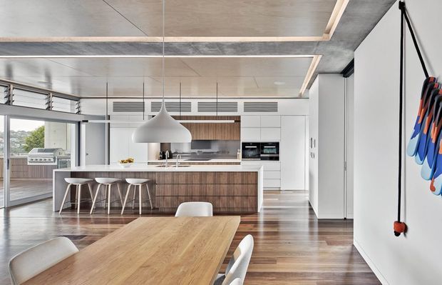Bright contemporary open-plan kitchen and dining area with a large timber island, integrated white cabinetry, pendant lighting, timber dining table, and expansive glazing opening toward an outdoor deck. The ceiling features timber-lined recessed panels and exposed concrete-like structural bands, while polished timber flooring runs throughout.