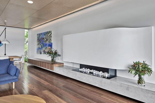 Contemporary living room with a long built-in concrete media console and a large white projection screen or concealed television feature. The room has dark timber flooring, a smooth white wall, timber veneer cabinetry, a linear recessed ceiling light, and a minimalist suspended pendant over a dining area. A blue upholstered armchair and restrained decorative greenery add colour and warmth.