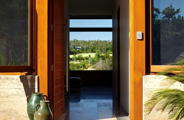 A sunlit exterior entry view looking through an open timber front door into a dark interior hallway or living space. The facade combines warm orange-brown timber door and window frames with pale textured rendered walls. Large screened windows flank the entrance, while a distant landscaped rural view is visible through the interior glazing. Decorative ceramic vessels sit beside the doorway and palm foliage appears at the edge of the frame.