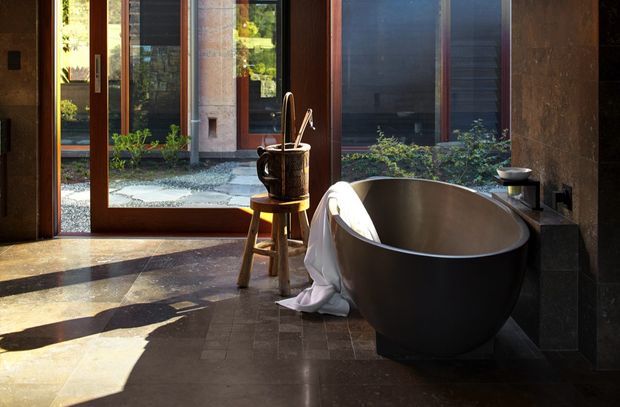 A sunlit, modern bathroom featuring a large freestanding oval bathtub beside dark stone or tile-clad walls. Floor-to-ceiling timber-framed glazing and a glass door open toward a private gravel courtyard with planting. A small timber stool with a dark vessel sits near the tub, while a dark stone-clad ledge or vanity and minimalist black fixtures line the right wall. Strong sunlight creates dramatic shadows across the stone floor.