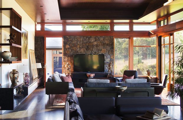 A spacious, light-filled living room with extensive timber-framed glazing, a dark timber ceiling, polished dark wood flooring, and a dramatic natural-stone feature wall surrounding a wall-mounted television. Multiple dark leather sofas and armchairs are arranged around the central seating area, with built-in display shelving and cabinetry along the left wall. The room has a warm, refined mid-century-inspired character and strong indoor-outdoor visual connection.