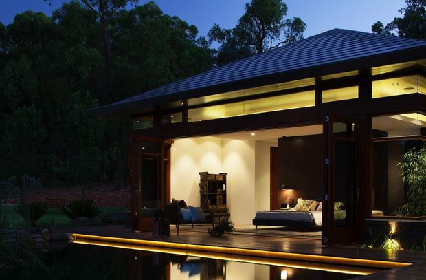 Twilight exterior view of a modern single-storey pavilion-style house set among mature trees. The building has a broad low-pitched dark metal roof, extensive floor-to-ceiling glazing, dark timber-framed openings, warm interior lighting, and a raised timber deck extending over a reflective pool or water feature. A bedroom and seating area are visible through the open glazing.
