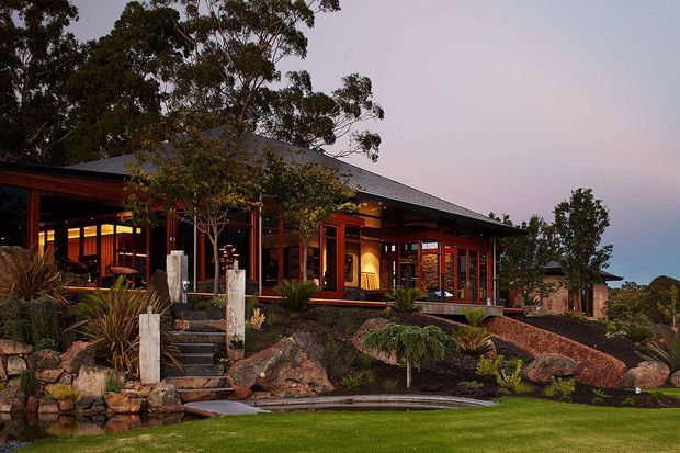 A low-slung, single-storey residence set within a landscaped garden at dusk. The home features extensive timber-framed glazing, warm interior lighting, dark grey pitched roofing, deep covered verandas, and a naturalistic rock garden with stone steps and water feature. Mature trees and layered planting surround the building.
