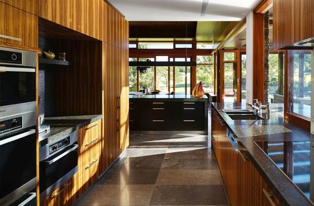 A spacious, highly detailed kitchen with extensive dark timber cabinetry and vertical wood panelling, integrated stainless-steel appliances, dark stone benchtops, and a central island. Large timber-framed windows and clerestory glazing bring in abundant natural light and overlook greenery. The room has a long galley-like layout with a polished, warm mid-century character.