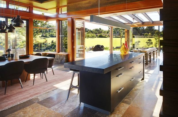 A spacious open-plan kitchen and dining area with a large dark island, integrated drawers, pale stone benchtop, timber-framed floor-to-ceiling glazing, and warm timber ceiling beams. A dining table sits to the left beneath pendant lights, while the glazed opening provides views across a landscaped rural property. The room is bright, warm, and strongly connected to the outdoors.
