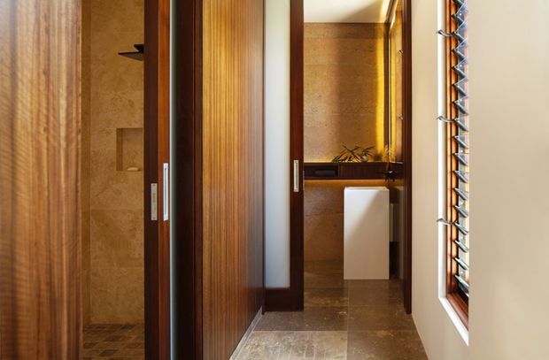 A warm, refined bathroom interior viewed through partially open timber doors. The space features rich vertical wood-panelled doors and trim, tan stone or tile wall surfaces, matching stone floor tiles, a floating white vanity, and a narrow louvred window providing filtered natural light. The composition suggests a compact ensuite or bathroom with a shower area visible to the left and vanity area at the rear.