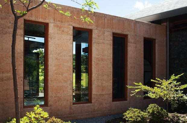 Exterior view of a contemporary building with a textured earthen-toned rendered wall, tall narrow dark-framed windows and doors, and a projecting corrugated metal roof. Landscaped planting sits in the foreground, while the glazing reflects or reveals greenery and an interior space.