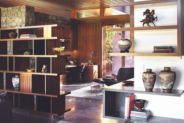 A richly detailed mid-century-inspired living room and study space with dark timber wall panelling, a stone feature wall, open room-divider shelving, and a sculptural built-in display unit. The foreground shelving separates the living area from a compact desk and lounge zone beyond. Warm timber-lined ceilings and walls, polished dark flooring, decorative ceramics, books, artwork, and leather seating create a refined, dramatic interior.