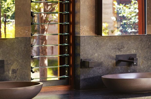 A dark, refined bathroom vanity scene with two sculptural vessel basins, wall-mounted rectangular faucets, a broad mirror, and a timber-framed window fitted with horizontal metal bars. The walls and countertop have a textured dark stone-like finish, with warm natural light and greenery visible outside.