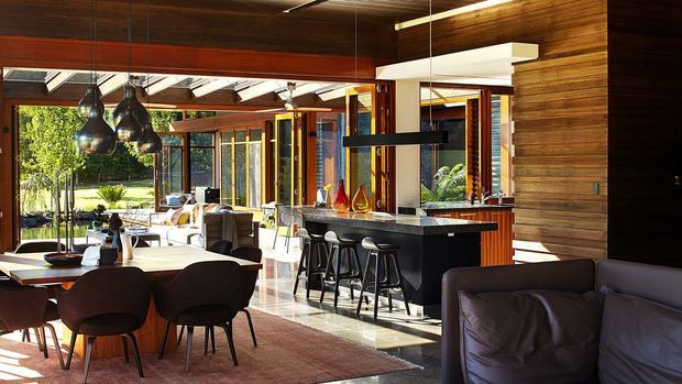 A spacious open-plan contemporary interior combining dining, living and kitchen areas. The room features extensive warm timber wall and ceiling lining, large glazed openings to a landscaped exterior, a dark stone kitchen island with integrated breakfast bar, pendant lighting, a dining table with upholstered chairs, and a leather sofa in the foreground. Strong indoor-outdoor connection and abundant natural light create a warm, refined atmosphere.