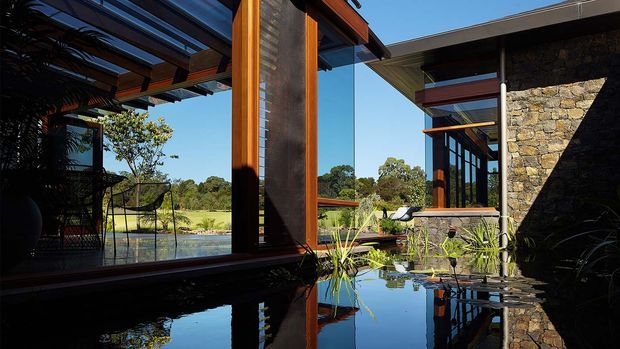 Exterior architectural view of a modern, low-profile residence arranged around a reflective water feature or pond. The composition highlights extensive floor-to-ceiling glazing, warm timber frames and pergola structures, dark metal rooflines, and a prominent rough natural-stone wall. Landscaped planting and an open rural-looking setting are visible beyond the building.