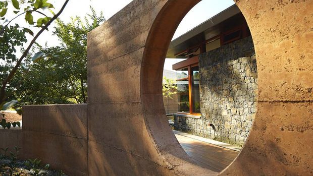 Exterior architectural view framed through a large sculptural circular arch opening in a warm earthen-toned wall. Beyond it is a courtyard or entry area with a dark natural stone masonry wall, timber decking, timber-framed glazing, and a low contemporary roof form, surrounded by mature greenery. The composition highlights the heavy curved masonry form and material contrast between rendered wall, rough stone, timber, and glass.