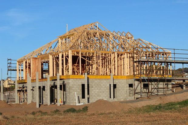 A partially constructed multi-level residential building on a dry, open site, showing concrete block lower walls, extensive timber wall and roof framing, scaffolding, and a pitched gable roof structure under a clear blue sky.