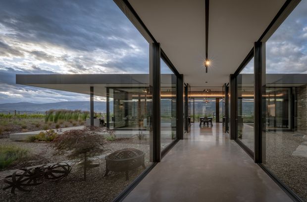 A dramatic contemporary glazed corridor or entry passage with floor-to-ceiling black-framed glass walls, a smooth concrete floor, and a slim linear ceiling with a pendant light. The passage leads toward a dining area and opens onto a sparsely landscaped rocky site with broad mountain views. Dark stone walls and exposed structural framing reinforce the modern architectural character.