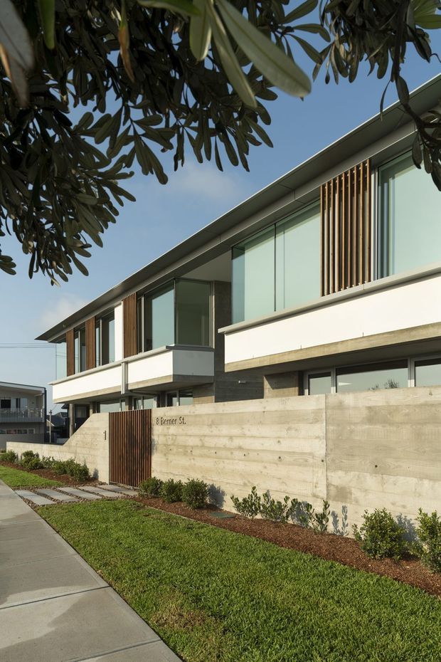 Street-level exterior view of a contemporary multi-storey residential building. The facade combines crisp white rendered bands, extensive floor-to-ceiling glazing, exposed concrete surfaces, and vertical timber screening. A concrete boundary wall and timber pedestrian gate front a landscaped lawn and paved footpath, while mature tree foliage frames the upper portion of the image.