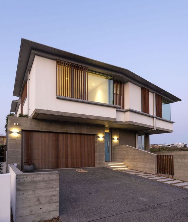 Exterior photograph of a modern two-storey residence at dusk, featuring a broad low-pitched roof, white rendered upper walls, exposed concrete walls and garage surround, timber vertical screening, a timber garage door, recessed warm lighting, and a sloping driveway with stepped pedestrian access.