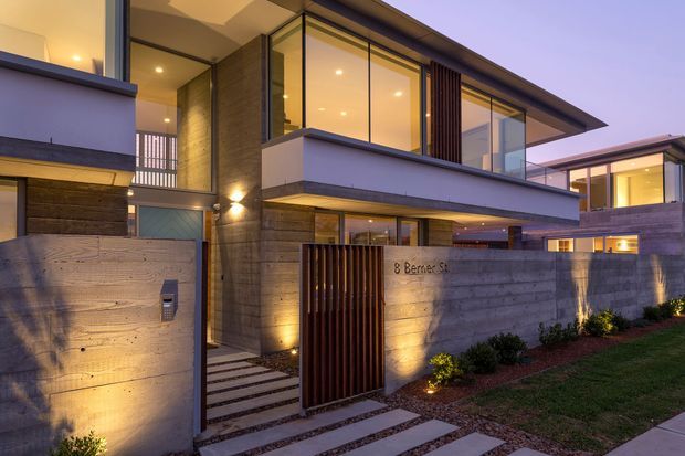 Twilight view of a contemporary multi-level residential facade with strong horizontal rooflines, concrete walls, large glazed openings, timber-look garage and entry elements, and a recessed pedestrian gate. Illuminated stepping-stone paving leads to the entrance, with low landscaped planting along the boundary wall.