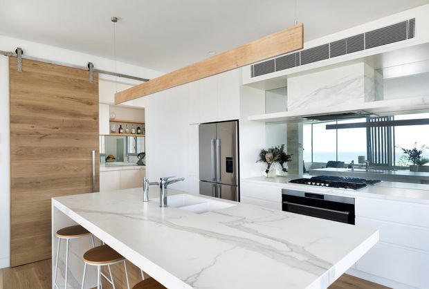 Bright contemporary kitchen with a large white marble-look island, integrated sink and two stools, white handleless cabinetry, stainless-steel refrigerator and built-in oven. A timber sliding barn door and timber overhead beam add warmth, while a mirrored backsplash reflects the cooktop and views through the glazing.