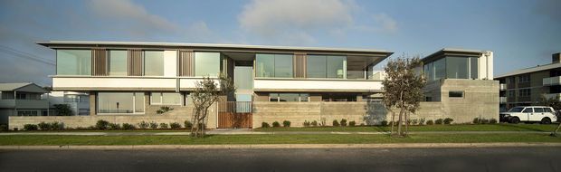Wide exterior view of a large contemporary two-storey residence with a long, low-profile form, projecting flat roof, extensive floor-to-ceiling glazing, pale rendered walls, and sections of exposed light masonry. The house fronts a landscaped verge and paved street, with neighbouring modern homes visible around it.