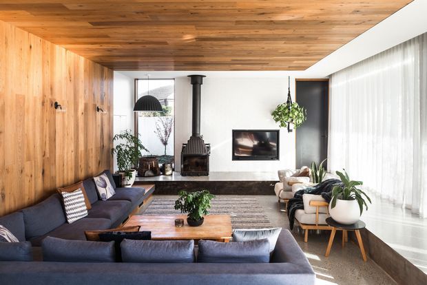 A warm, contemporary living room centered around a dark sectional sofa and timber coffee table. The room features extensive natural timber wall and ceiling lining, a freestanding black wood-burning stove, a dark stone or concrete fireplace hearth, white walls, floor-to-ceiling sheer curtains, indoor plants, and a large area rug. Glazing and a bright adjoining area are visible at the rear, while the overall composition emphasizes the living space and its fireplace.