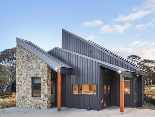 Exterior view of a contemporary single-storey building with an angular pitched roof and dark vertical corrugated metal cladding. A prominent natural irregular-stone wall forms the left wing, with timber posts and recessed glazed entry areas. The structure sits in a lightly wooded rural setting with a concrete approach and clear blue sky.