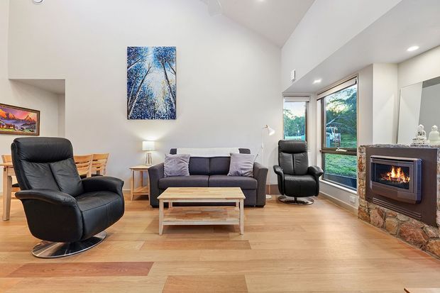 Bright, open-plan living room with a high sloped ceiling, pale timber flooring, a dark sofa and leather lounge chairs, and a stone-clad fireplace with a built-in firebox. A dining area is visible at the left, while large windows overlook greenery. The room has a clean contemporary feel with warm timber accents and abundant natural light.