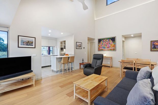 Bright open-plan living area with light timber flooring, a dark sectional sofa, timber coffee table, dining table and chairs, and a leather recliner. The space opens directly to a compact white kitchen with a peninsula and bar stools. High ceilings, clerestory glazing, recessed lighting, and a large colourful artwork create an airy contemporary interior.