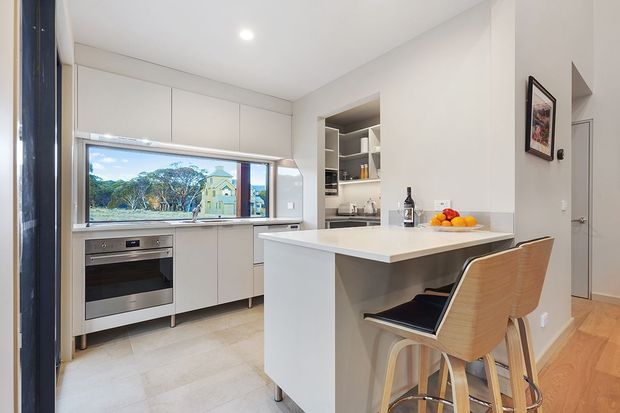 Bright contemporary kitchen with a compact island and two bar stools, light cabinetry, integrated oven, pale stone-look flooring, and a wide window overlooking greenery and neighbouring houses. A recessed open shelf and sink area sit behind the island, with timber flooring extending into the adjacent space.
