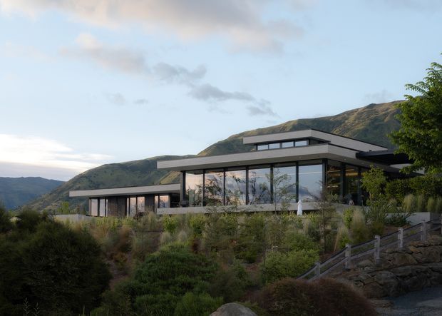 A contemporary single-storey hillside residence with broad floor-to-ceiling glazing, projecting flat roof planes, dark horizontal framing, and low rectilinear volumes. The house is integrated into a landscaped, sloping site with native-looking planting, stone retaining walls, and steps, backed by rugged mountains. The image is an exterior architectural view in soft natural light.